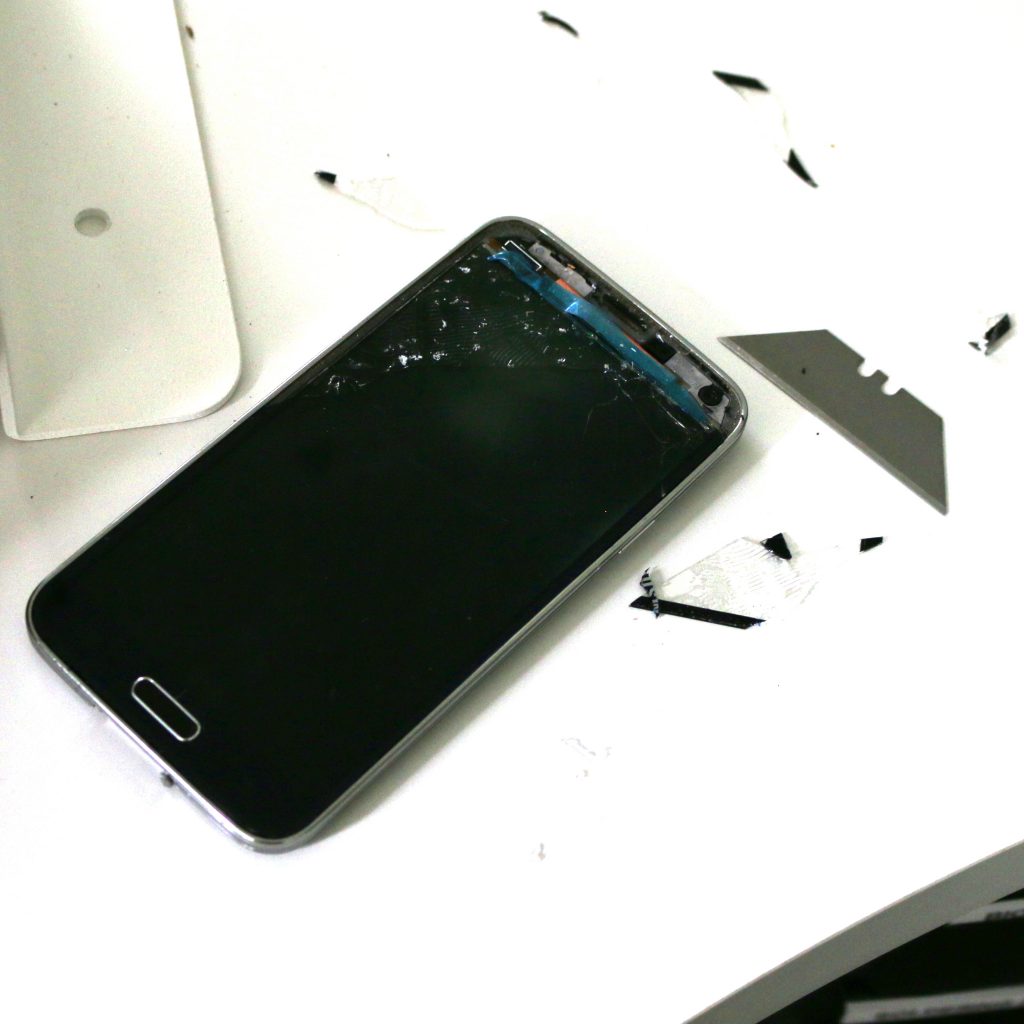 A Samsung Galaxy S5 with its screen removed, showing a cracked, shattered LCD panel and adhesive debris on a white table next to a razor blade.