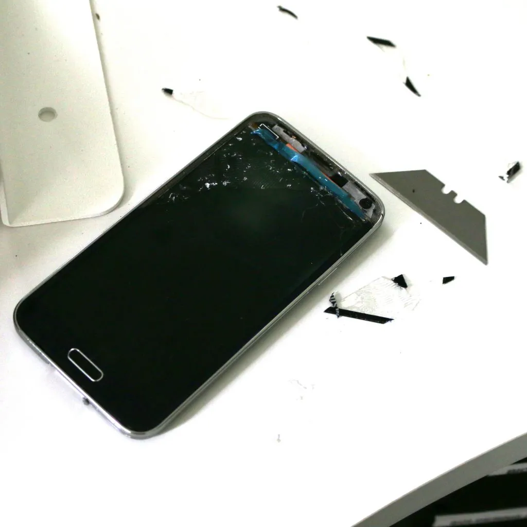 A Samsung Galaxy S5 with its screen removed, showing a cracked, shattered LCD panel and adhesive debris on a white table next to a razor blade.