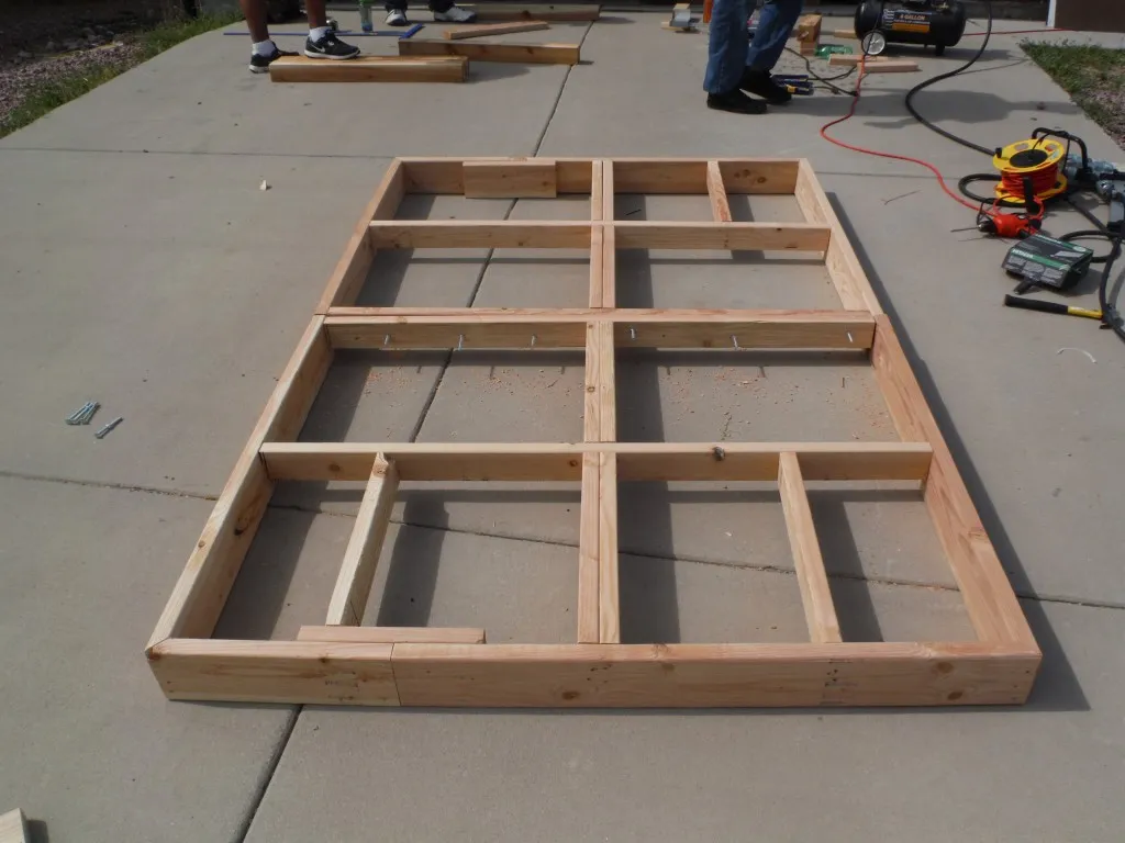 A wooden frame for the robot arena tabletop assembled on a driveway, with two people working in the background near an air compressor.