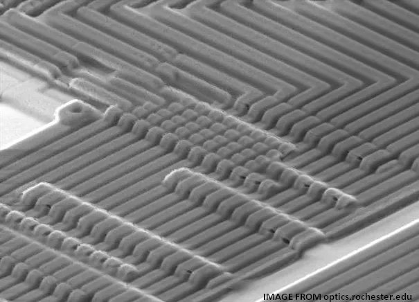 A grayscale electron microscope scan of aluminum trace patterns on a silicon chip, credited to optics.rochester.edu.